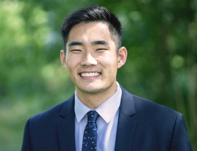 Image description: Zach is an Asian man with short black hair in a crew cut. He is wearing a navy suit and tie with flowers on it and a lavender dress shirt. There is a blurred green background of nature foliage as well.